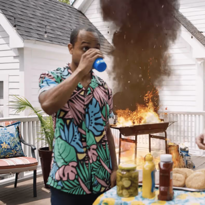 Man in colorful tropical shirt drinking from a blue cup while a barbecue grill behind him is engulfed in flames and thick black smoke on a patio.
