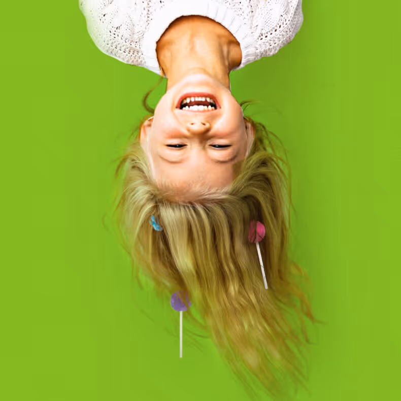 Upside-down young girl with blonde hair decorated with colorful lollipops, smiling against a bright green background.