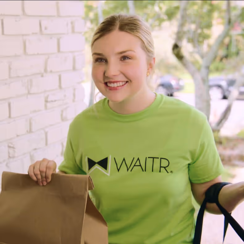 Smiling delivery woman wearing a green WAITR shirt holding a brown paper bag and a black insulated bag at a doorway.