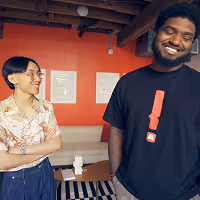 Khoi and Justin smiling and standing indoors against a red wall with framed pictures and wooden ceiling beams.